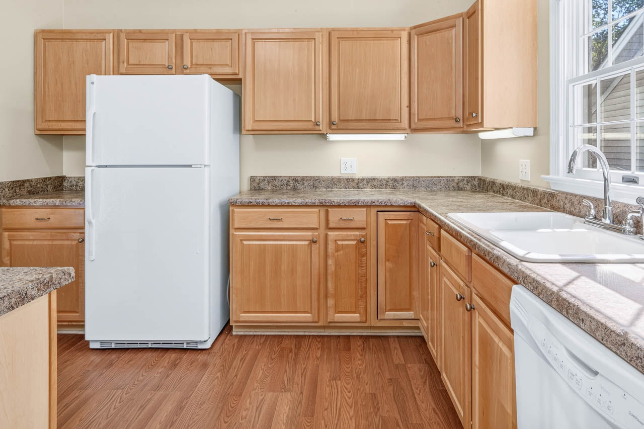 A kitchen with light wood cabinets, white appliances including a refrigerator and dishwasher, brown marble countertops, a double sink by a window, and wood flooring.