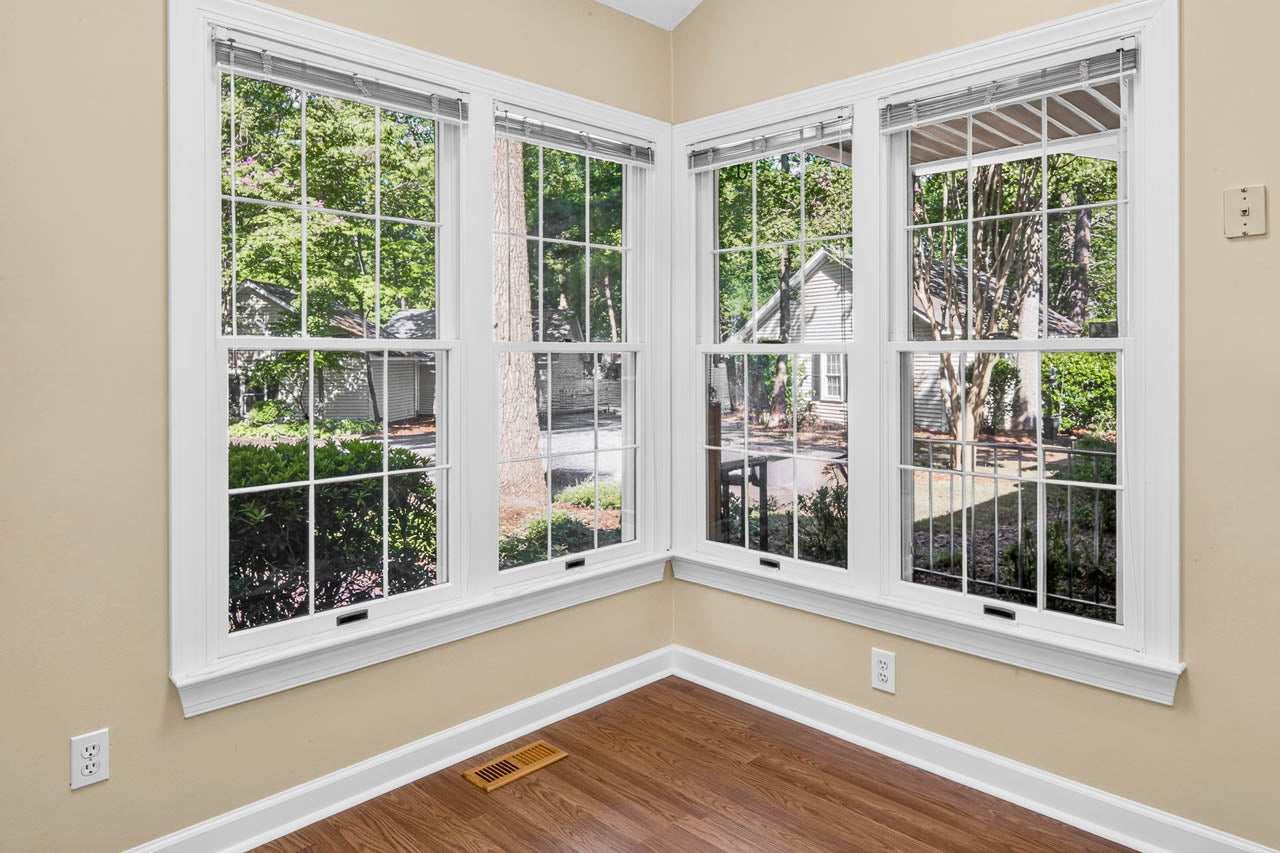 Corner of a room with light beige walls, large white-trimmed windows, and wood flooring. View of trees, greenery, and a house outside through the windows.