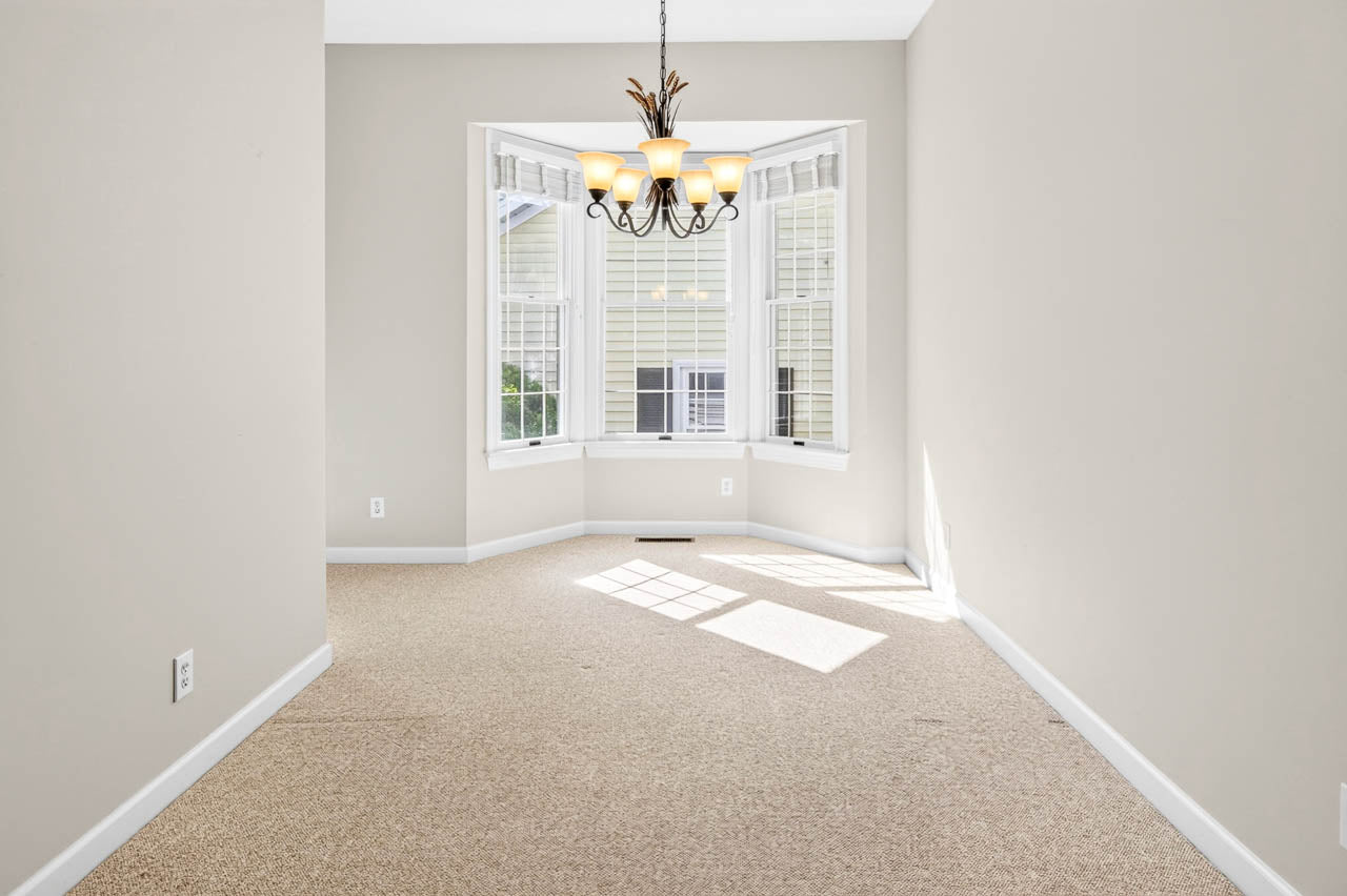 Empty, carpeted room with beige walls, a hanging chandelier, and a large bay window with white shutters letting in sunlight.