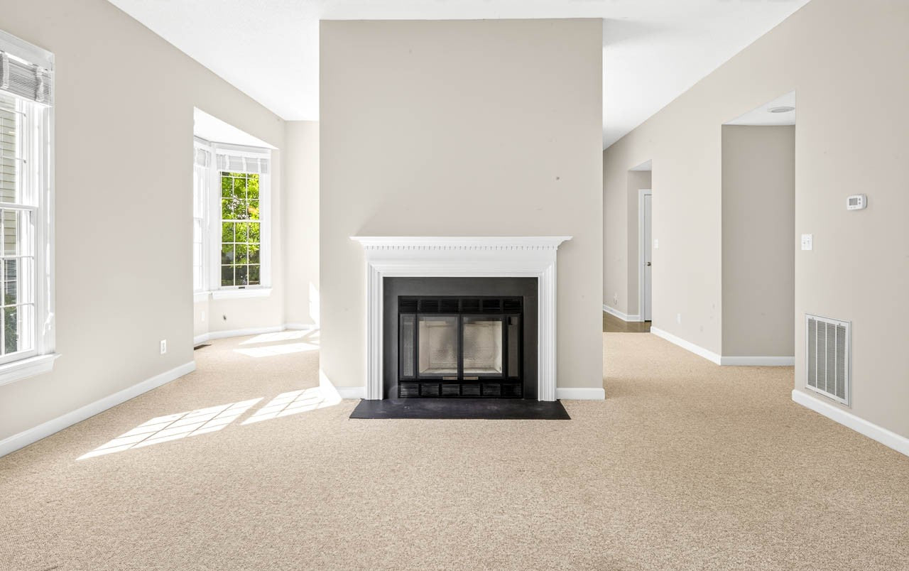 Bright, empty living room with beige walls and carpet, white trim, a central fireplace with a black hearth, and large windows letting in natural light.