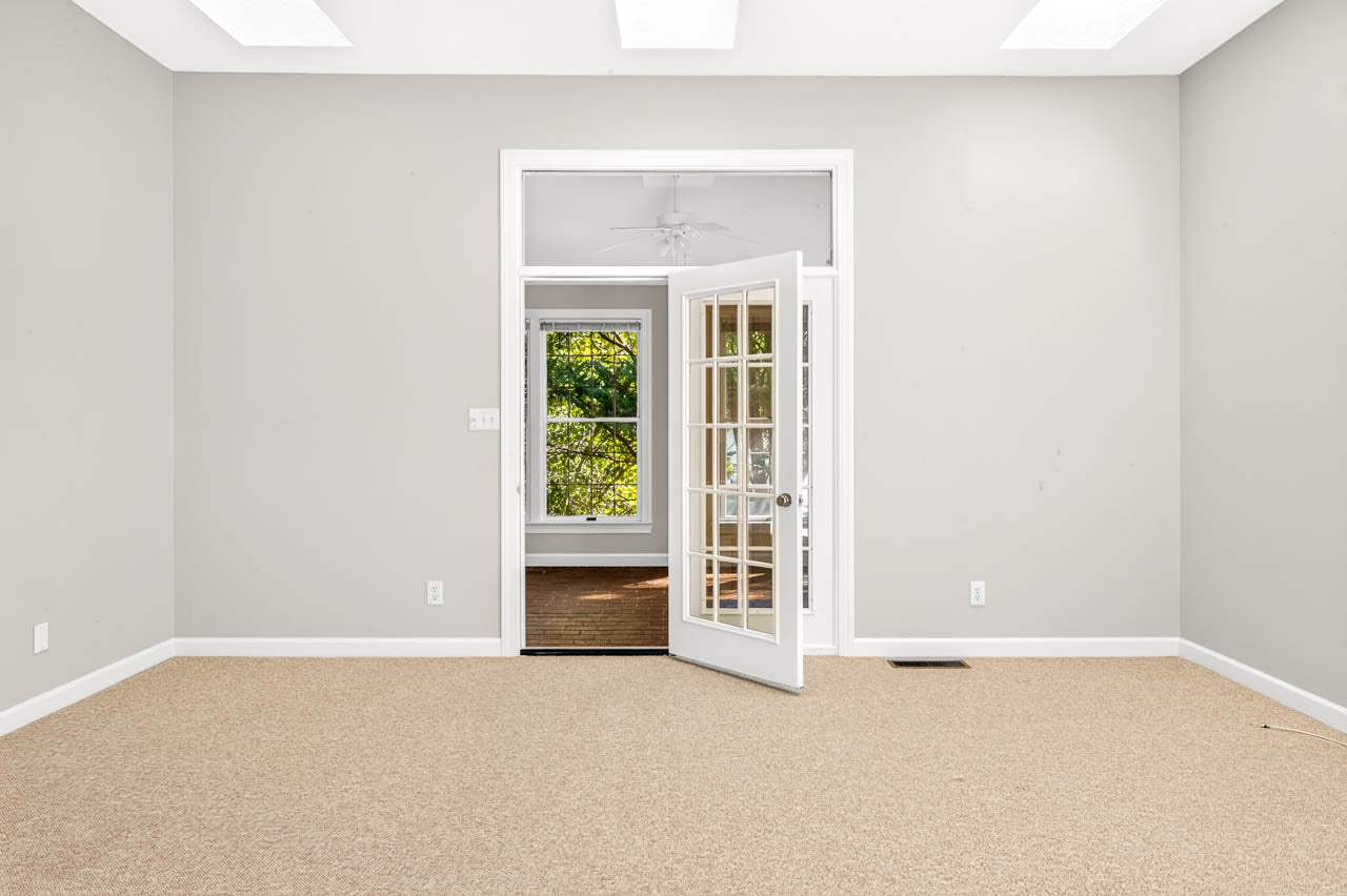 Empty beige-carpeted room with light gray walls, white trim, and French doors leading to a sunlit room with wood flooring and large windows showing green trees outside.