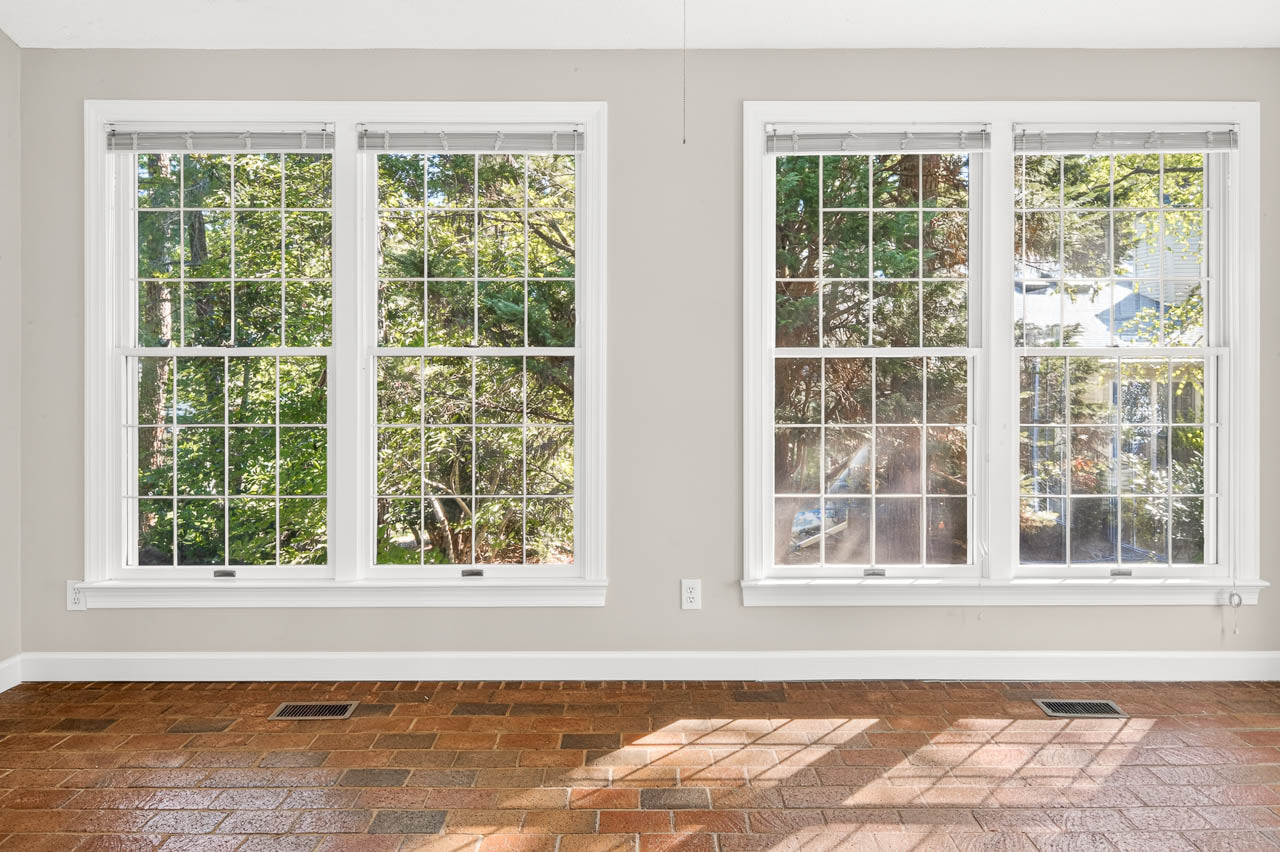 Two large windows with white frames let in natural light, showcasing a view of green trees outside. The room has light gray walls and a reddish-brown tiled floor.