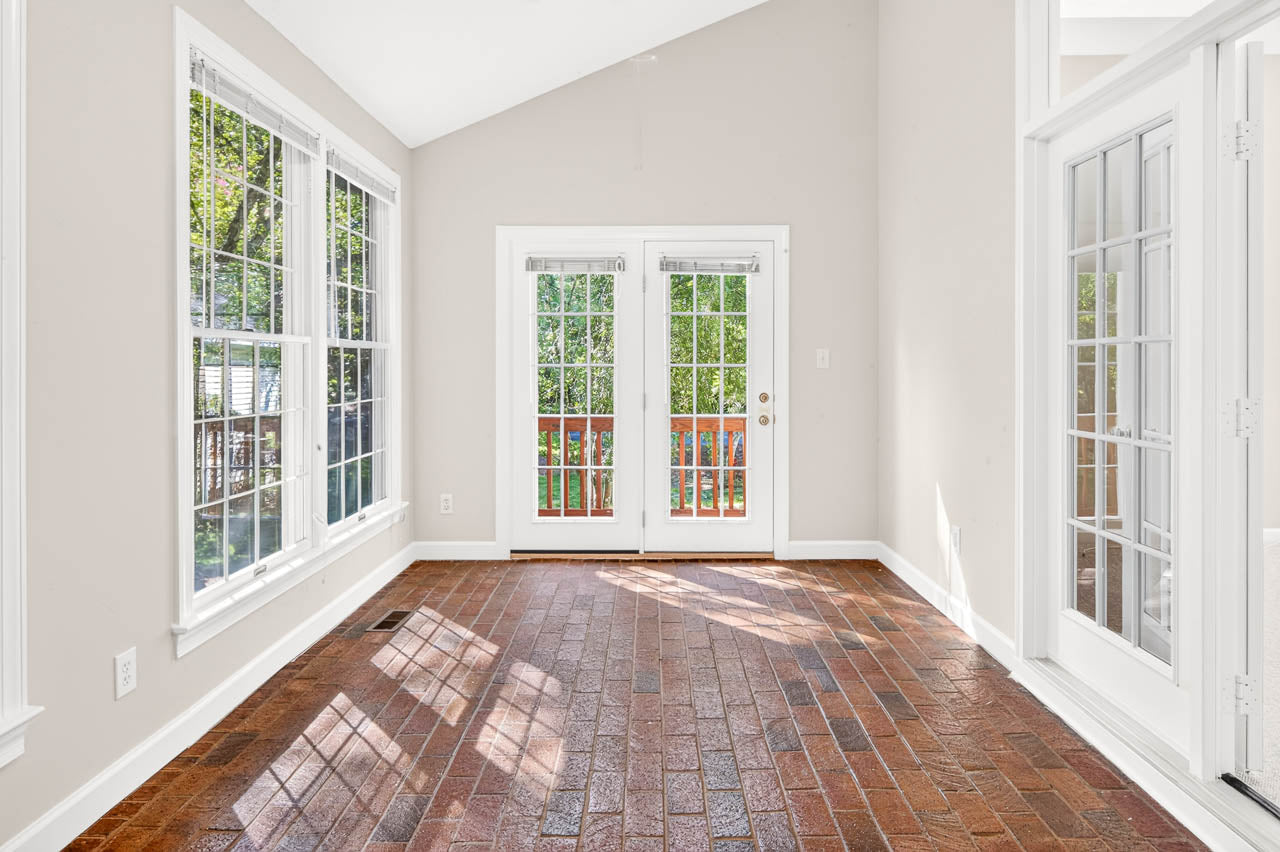 Bright sunroom with large windows, French doors, and red brick flooring. Sunlight streams in, casting shadows across the floor. The room has white trim and neutral walls.