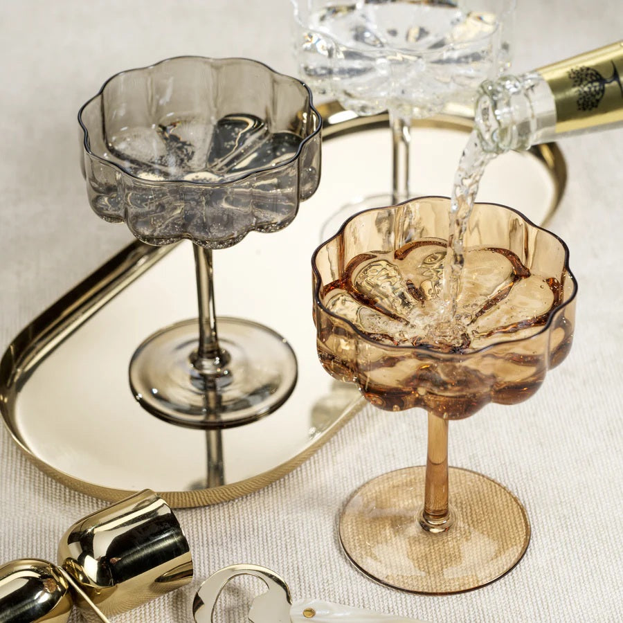 The ROCCO HANDBLOWN COUPE - AMBER by ZODAX features a brown-tinted, handblown amber glass with a scalloped, fluted bowl and a long stem, standing on a round base against a white background.