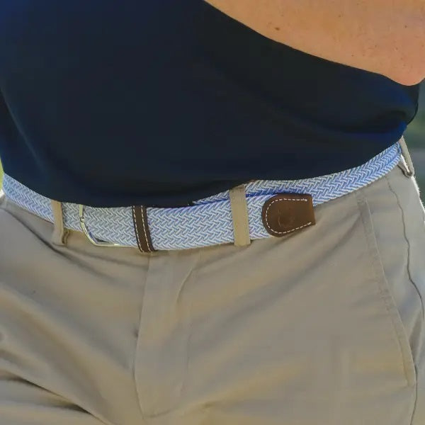 The ROOSTAS - SARASOTA WOVEN STRETCH BELT by ROOSTAS features sky blue and white weaving, brown leather ends, and a silver buckle, shown coiled with the buckle at the front.