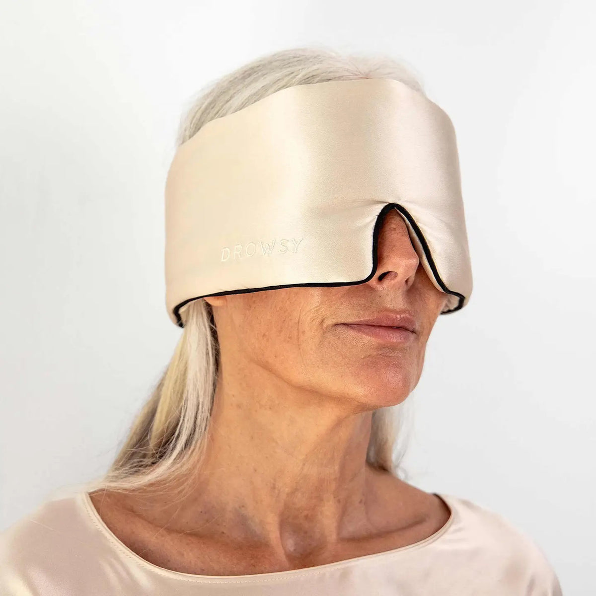 A person wearing a white tank top is lying on a pillow with the DROWSY - SATIN SLEEP MASK by DROWSY SLEEP CO covering their eyes. They look relaxed, enjoying beauty sleep as their hand rests gently on the pillow.