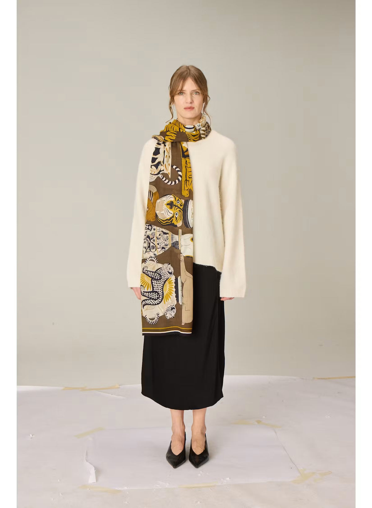 The INOUI EDITIONS - COLLECTIONNEUR Wool Scarf with Cats in Khaki by INOUI is a rectangular accessory crafted in India from 100% wool. It features an artistic collage-like pattern of illustrated cats and circular motifs in shades of gold, white, black, and gray set against a dark khaki background. Measuring 70 x 190 cm, this striking scarf adds both warmth and a touch of artistry to any ensemble.