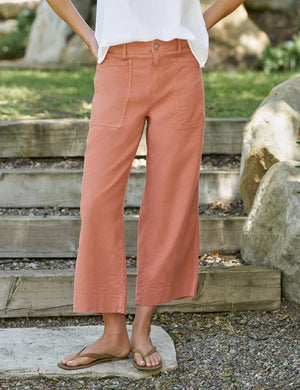 A person wearing the FRANK & EILEEN Limerick Cropped Wide Leg Carpenter Pant in Italian Performance Linen Canyon, paired with a white top and tan sandals, stands on outdoor stone steps with hands in pockets; the background features grass, rocks, and sunlight.