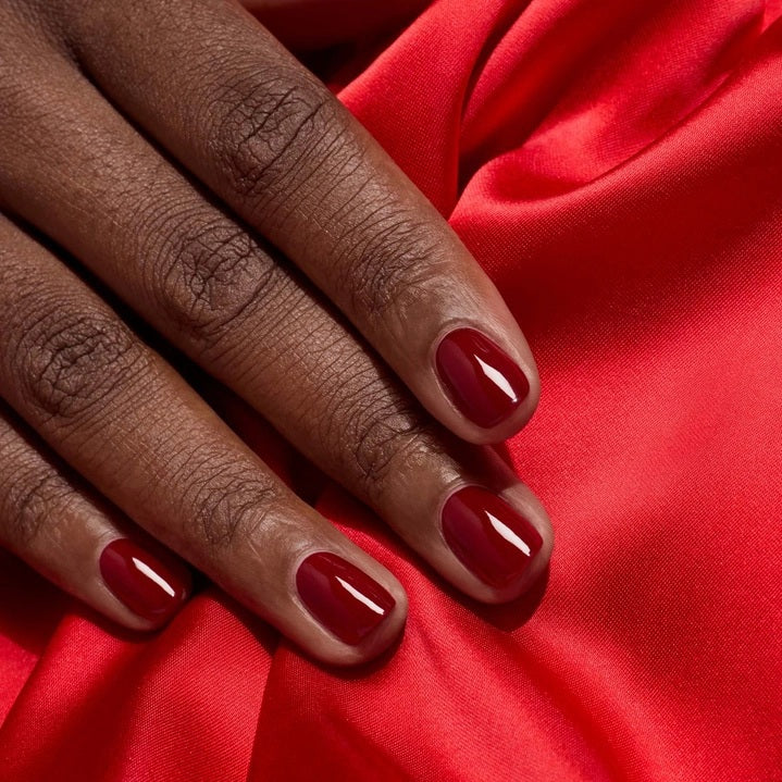 A hand with neatly manicured nails painted in a rich shade of dark red rests on red fabric beside a matching bottle of MANUCURIST INC’s GREEN™ Nail Polish in Dark Pansy, a non-toxic and vegan formula.