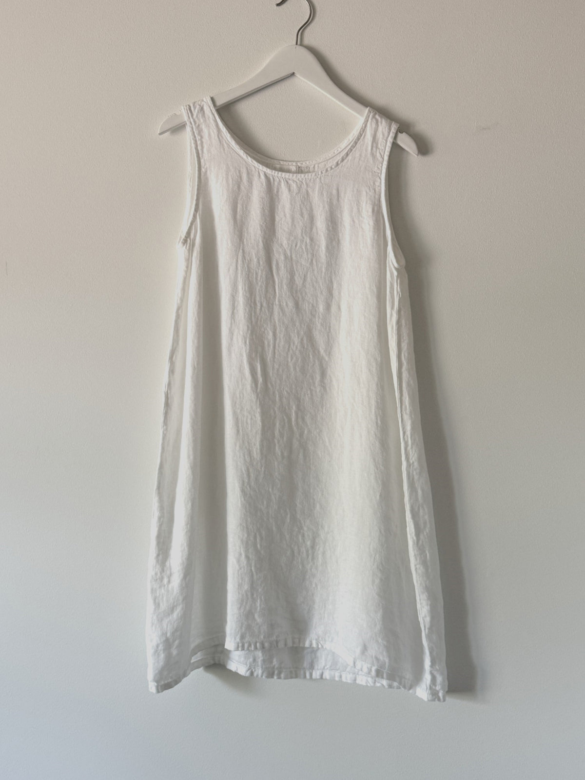 The CP SHADES - JESS SLEEVELESS DRESS, an A-line linen dress, hangs on a white clothes hanger against a light-colored wall. The dress features a round neckline and a minimalist design, with its slightly wrinkled texture adding to its charm. This eco-friendly piece by CP SHADES showcases solar-powered production at its finest.