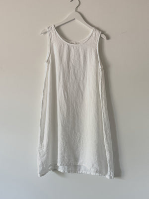 The CP SHADES - JESS SLEEVELESS DRESS, an A-line linen dress, hangs on a white clothes hanger against a light-colored wall. The dress features a round neckline and a minimalist design, with its slightly wrinkled texture adding to its charm. This eco-friendly piece by CP SHADES showcases solar-powered production at its finest.