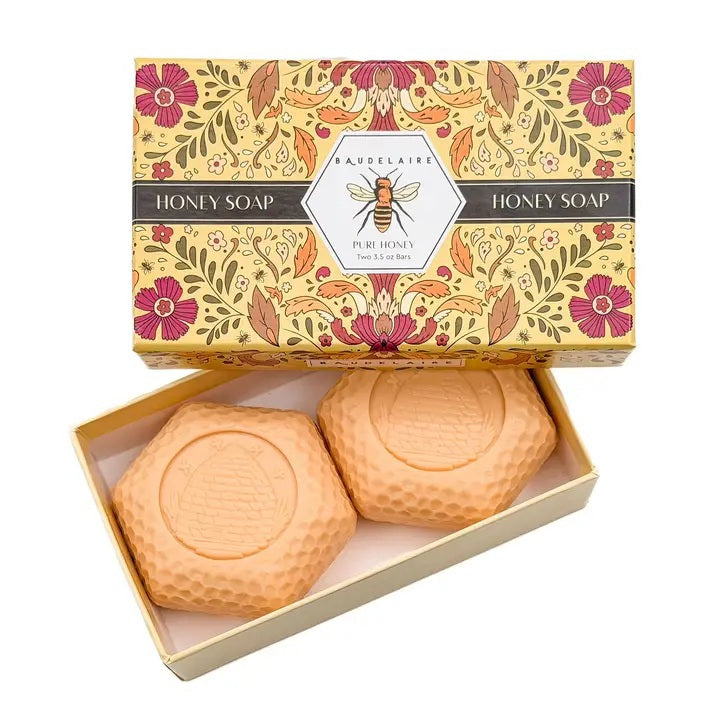 A floral box labeled “BAUDELAIRE - PURE HONEY SOAP 3.5 OZ, 2 BAR GIFT BOX” sits beside two honeycomb-shaped BAUDELAIRE Pure Honey Soaps featuring a bee design—each a gentle, triple-milled soap ideal for sensitive skin—accompanied by a white tray, yellow flowers, and stones on a white surface.