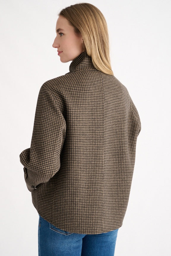 A woman stands against a plain white background wearing the KINROSS CASHMERE Houndstooth Button Mock Coat by KINROSS, featuring a brown and black houndstooth pattern, a high mock neckline, and front buttons, paired with blue jeans.