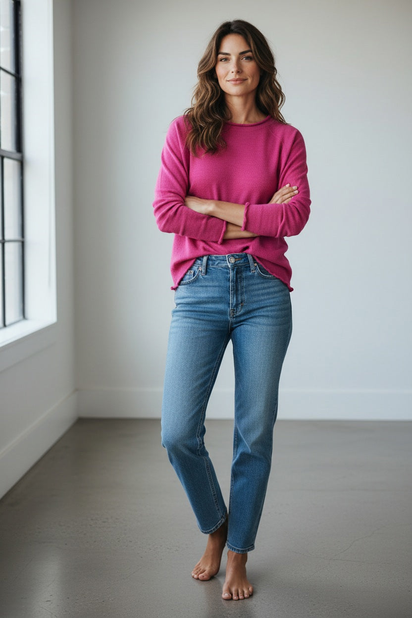 A person with short blonde hair stands in a doorway, arms crossed, wearing the MERSEA Catalina Ruffle Tee in Rapture Rose layered over a white blouse with blue jeans. Green leaves frame the foreground.