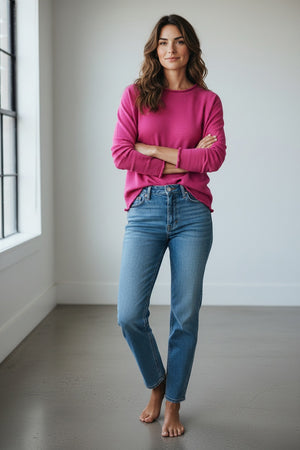 A person with short blonde hair stands in a doorway, arms crossed, wearing the MERSEA Catalina Ruffle Tee in Rapture Rose layered over a white blouse with blue jeans. Green leaves frame the foreground.