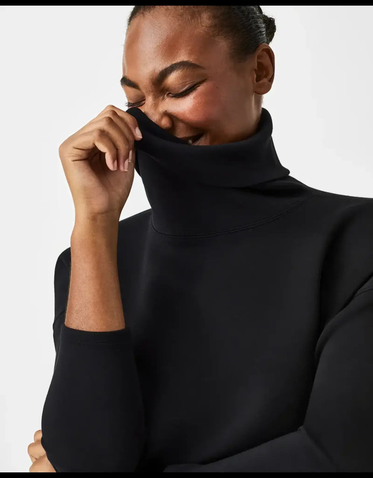 A woman wearing the SPANX AIRESSENTIALS Turtleneck Tunic in Black—a long-sleeved, relaxed fit sweater—stands against a plain white background, looking down and gently touching her arm.