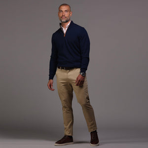 A man with short hair and a beard stands against a gray background wearing the COLLARS AND CO Navy with Brown Suede Trim Cashmere Touch Australian Merino Quarter Zip Sweater over a white shirt, khaki pants, and brown high-top sneakers, one hand in his pocket.