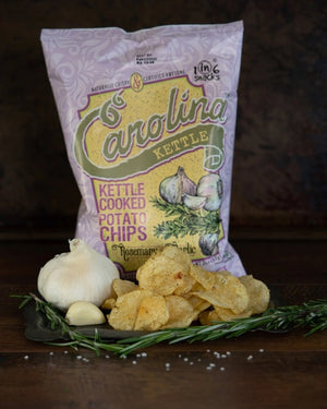 A 5 oz (142g) bag of 1 in 6 Snacks' Carolina Kettle Rosemary & Garlic Chips, bursting with garlic seasoning and fresh rosemary. The purple bag features images of both ingredients and labels indicating that these kettle chips are non-GMO, gluten-free, and naturally crispy.