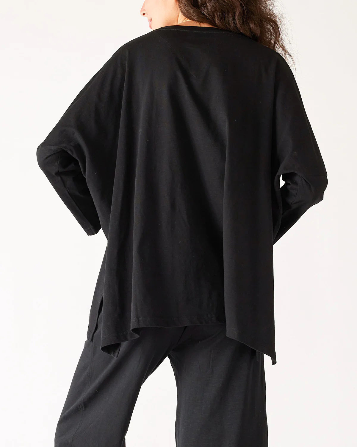 A woman with wavy brown hair models the MERSEA - CATALINA SLUB TEE—an oversized, long-sleeved white top with thin gray stripes—by MERSEA. She pairs it with blue jeans and stands against a plain, light background, hands behind her back.