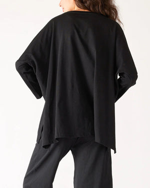 A woman with wavy brown hair models the MERSEA - CATALINA SLUB TEE—an oversized, long-sleeved white top with thin gray stripes—by MERSEA. She pairs it with blue jeans and stands against a plain, light background, hands behind her back.