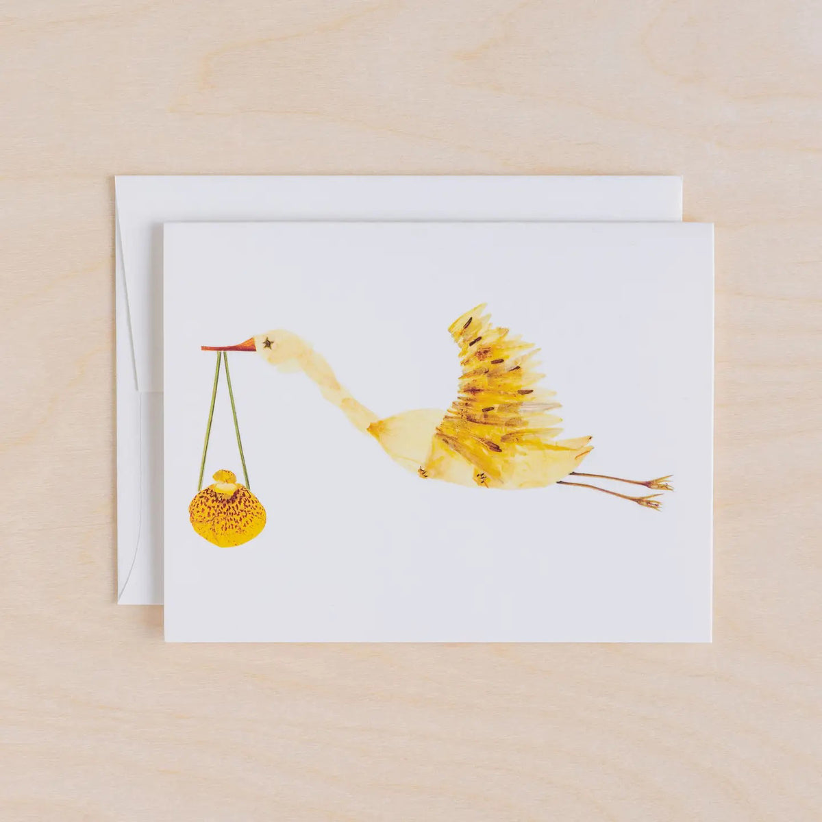 The PETAL PEOPLE PRESS - STORK CARD by PETAL PEOPLE PRESS features a yellow painted stork with a bundle, displayed on a white surface with green leaves, scissors, and a dish with a paperclip.