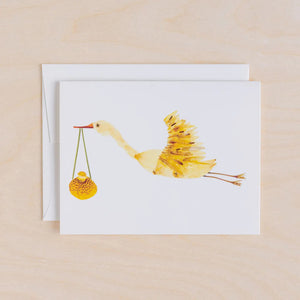 The PETAL PEOPLE PRESS - STORK CARD by PETAL PEOPLE PRESS features a yellow painted stork with a bundle, displayed on a white surface with green leaves, scissors, and a dish with a paperclip.