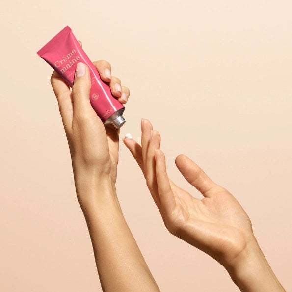 Close-up of two hands with light skin; one is gently applying MANUCURIST - COSMOS ORGANIC HAND CREAM, ORANGE BLOSSOM by MANUCURIST INC to the back of the other against a neutral beige background. Ideal for hand and nail care, this organic moisturizer is formulated with natural ingredients to leave fingers nourished. The nails are perfectly groomed and finished with a subtle pale nude polish for an elegant touch.
