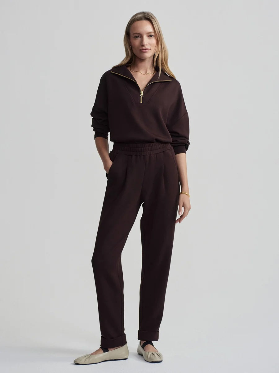 A person seen from behind wearing VARLEY - ROLLED CUFF PANT 25” IN COFFEE BEAN dark brown trousers with rolled cuffs and cream-colored ballet flats.