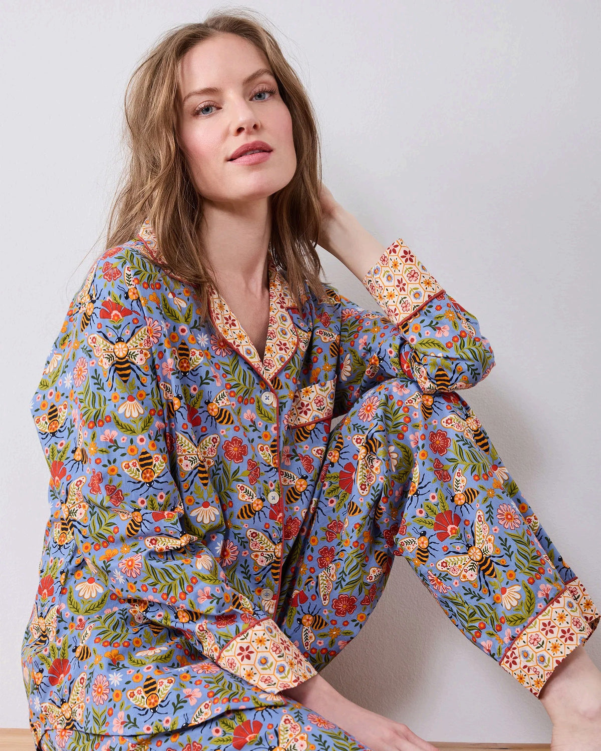 A woman with light brown hair sits against a white wall, wearing the PRINTFRESH - Bee Tapestry Long Pajama Set in Delphinium Blue. This organic cotton pajama set from PRINTFRESH features a colorful floral and bee pattern with vibrant red and yellow cuffs, collar, and pocket trim, beautifully blending playful design with cozy comfort.