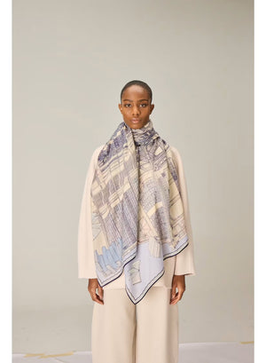 A person wearing a cream-colored outfit stands facing forward, draped in the INOUI EDITIONS VIVIENNE Wool Scarf in Grey—a large, patterned scarf featuring blue and beige tones. Made from 100% wool and crafted in India by INOUI, the scarf measures 100 x 190 cm and is set against a plain, light-colored background.