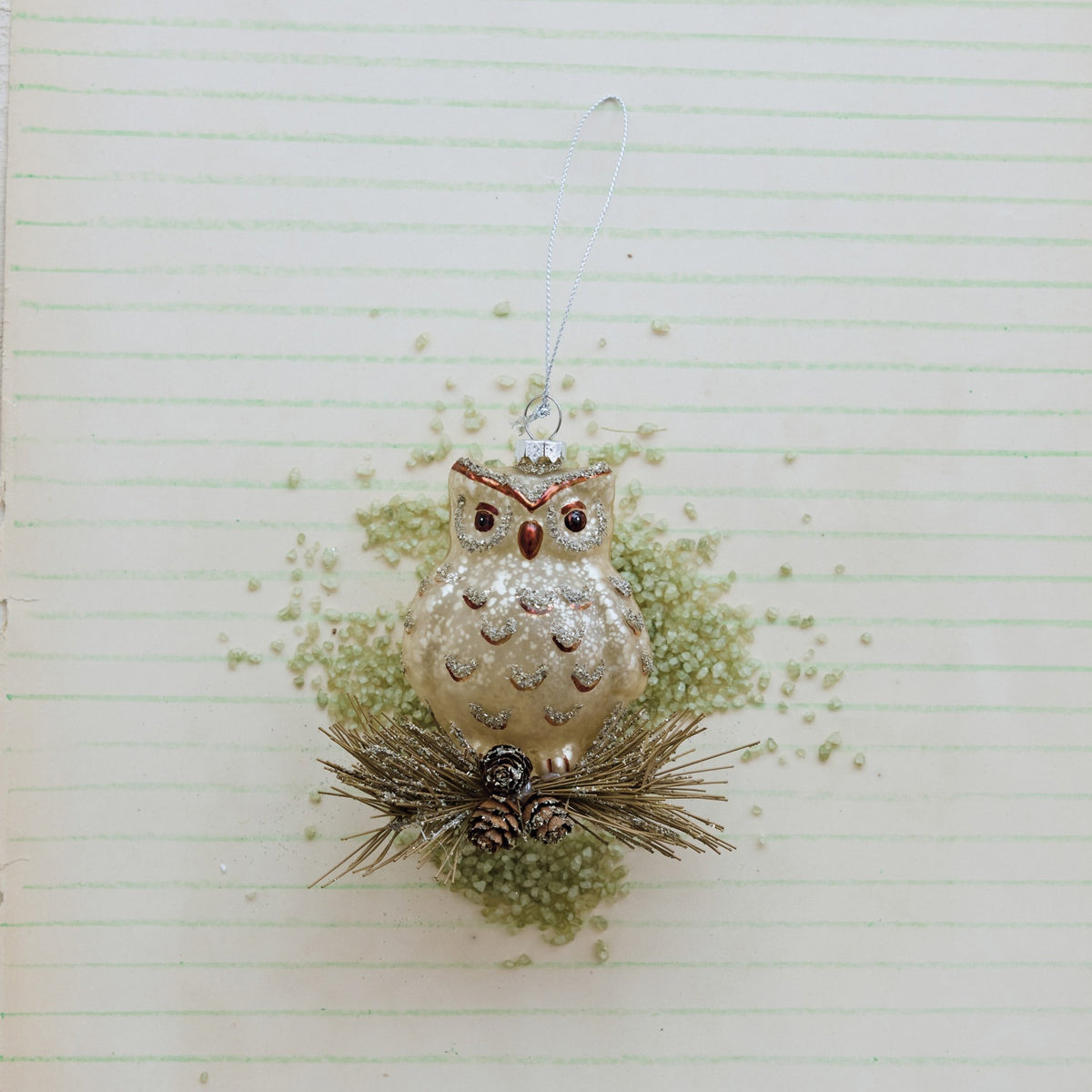 A GLASS OWL ORNAMENT WITH GLITTER perched on a cluster of pine needles and small glitter pinecones.