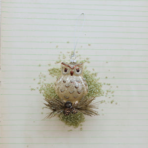 A GLASS OWL ORNAMENT WITH GLITTER perched on a cluster of pine needles and small glitter pinecones.