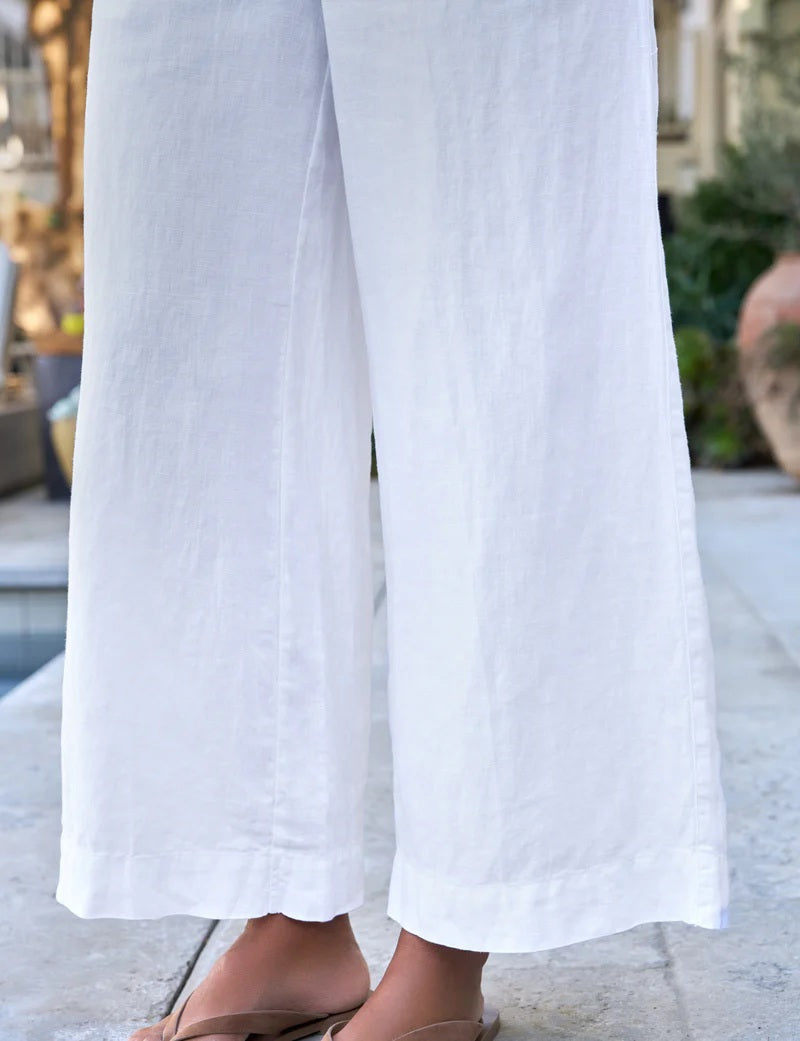 A person stands outdoors among plants and décor, hands in pockets, wearing FRANK & EILEEN’s Maisie Wide Leg Pull On Linen Pant in White with a light linen shirt and beige sandals.