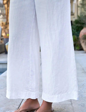 A person stands outdoors among plants and décor, hands in pockets, wearing FRANK & EILEEN’s Maisie Wide Leg Pull On Linen Pant in White with a light linen shirt and beige sandals.