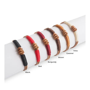 A person with a hand in their navy blue pants pocket wears two bracelets—one thin and dark, the other a thicker MAKARLA Men's Knot Leather Bracelet Adjustable in Caramel—set against a light background.