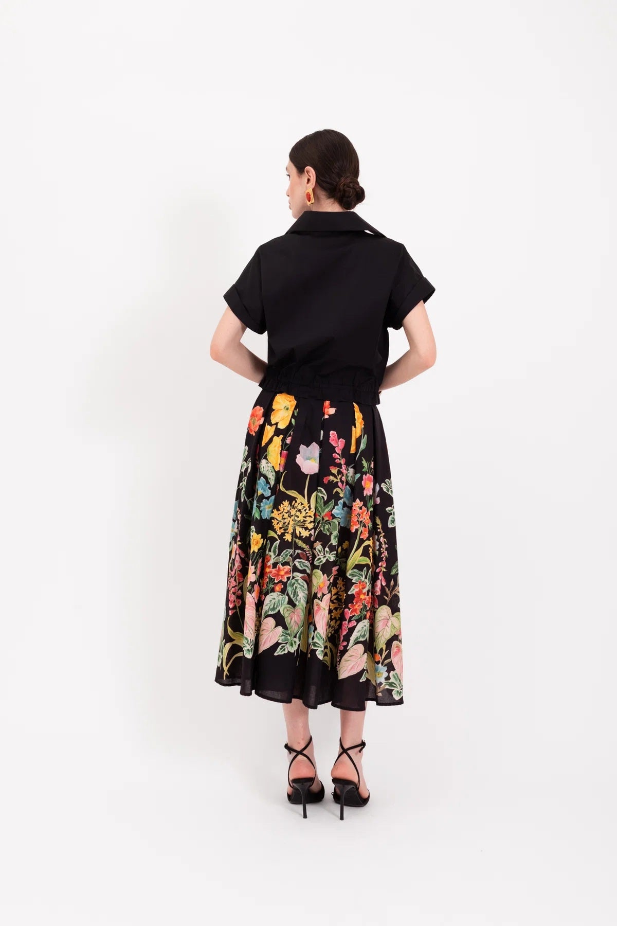 A woman stands against a white background wearing a black short-sleeve shirt paired with the LAVI - IRIS Botanical Skirt in Black Floral from LAVI, featuring colorful floral patterns, and completes the look with black high-heeled sandals.
