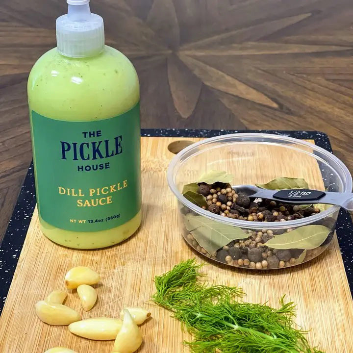 A 9.5-oz squeeze bottle of THE PICKLE HOUSE brand DILL PICKLE SAUCE, with a green label and white cap, sits on a light gray textured background. This Dill Garlic sauce is perfect as a marinade or salad dressing.