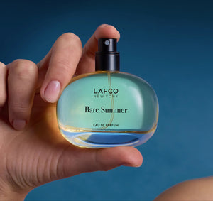 The LAFCO - FINE FRAGRANCE, BARE SUMMER 50ML perfume bottle, featuring a blue cap and displayed alongside delicate grasses, showcases its fresh and uplifting scent inspired by the sea and sand dunes. The text highlights opening, heart, and base fragrance notes that evoke the essence of summer. This vegan scent from LAFCO offers a clean, natural allure perfect for warm days.