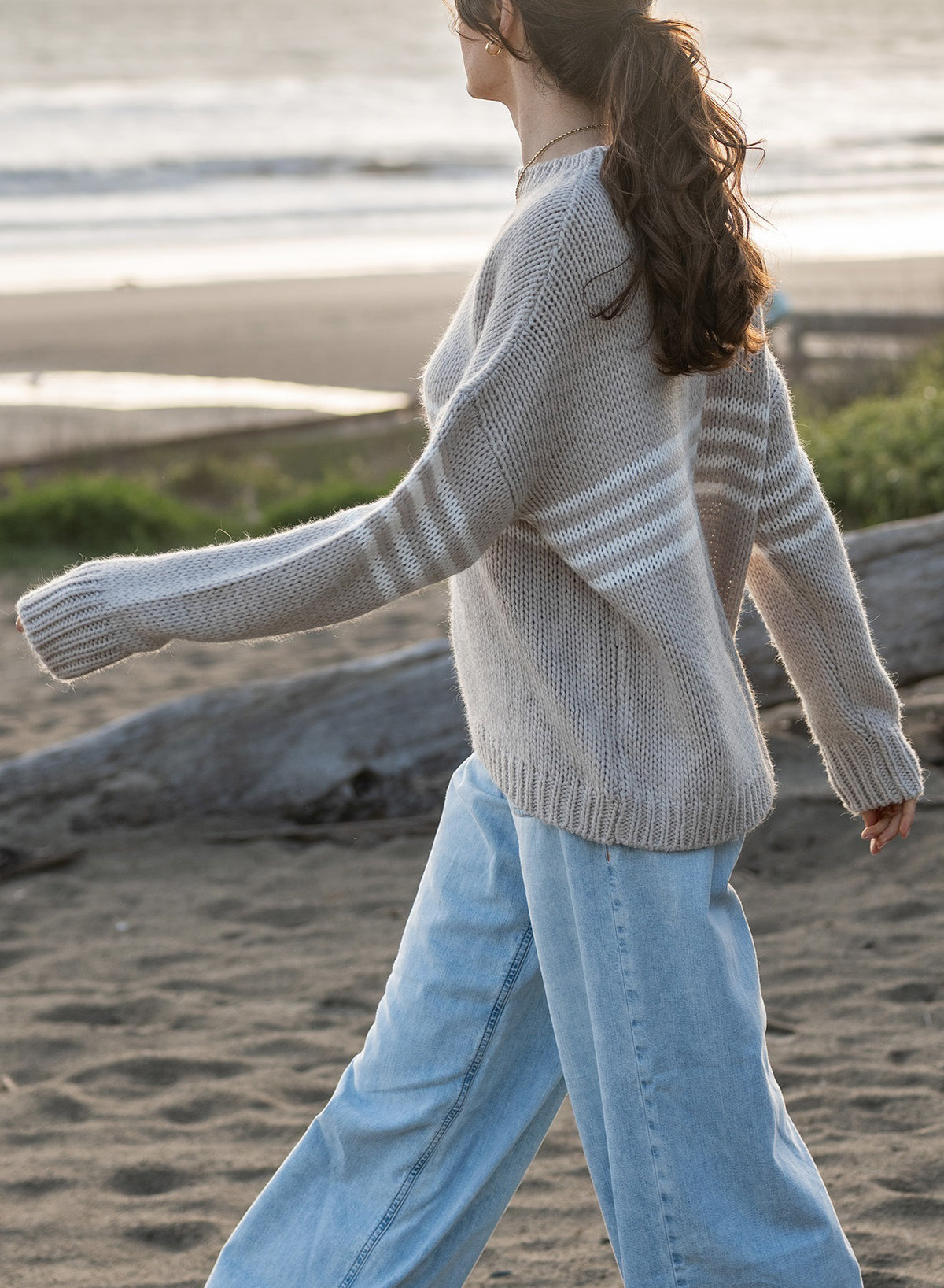 A woman with brown hair in a ponytail strolls a sandy beach at sunset, wearing the MERSEA - SEACOAST STRIPE SWEATER by MERSEA and light blue jeans as the sun glows over the ocean in the background.