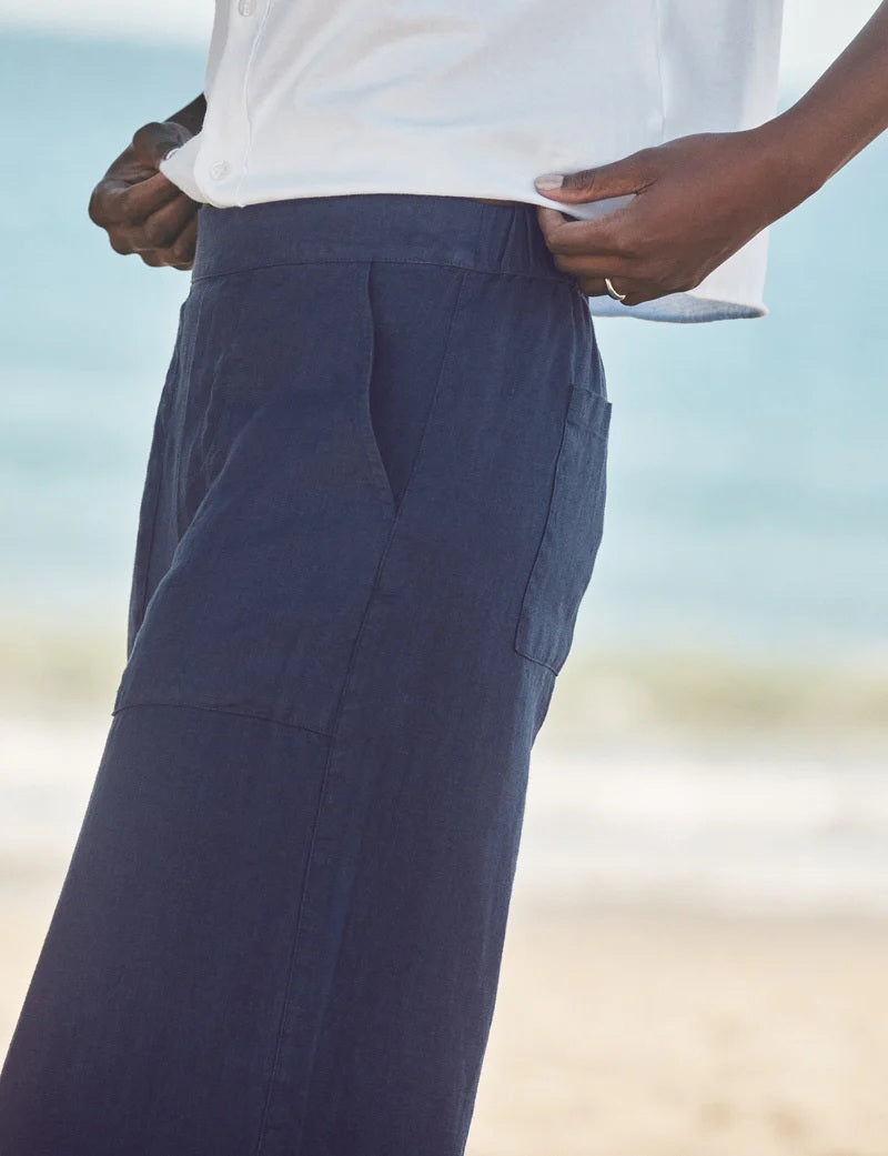 A person stands on a sandy beach wearing a white long-sleeve shirt, FRANK &amp; EILEEN MADELINE Patch Pocket Pull On Wide Leg Linen Pant in Navy, and light-colored sandals. The ocean is visible in the background.