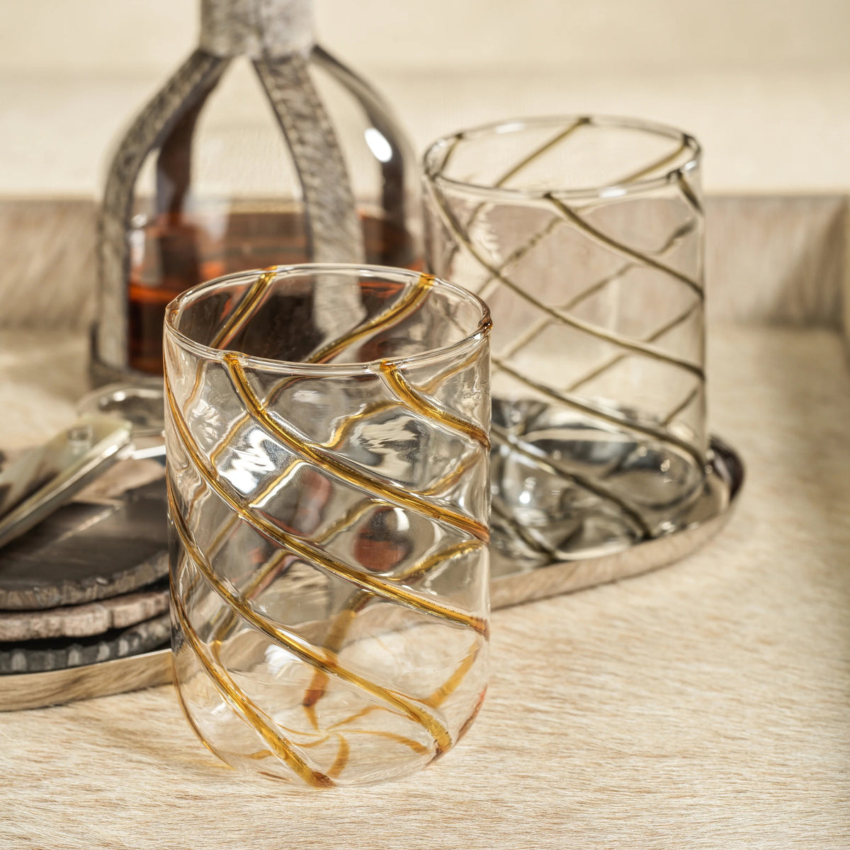 The AMBER SWIRL GLASS TUMBLER by ZODAX, a clear cylindrical glass adorned with bold gold diagonal swirl stripes, is elegantly displayed on a white background.