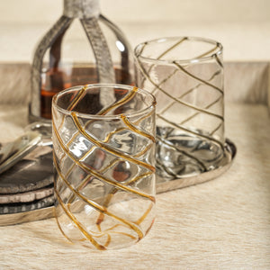 The AMBER SWIRL GLASS TUMBLER by ZODAX, a clear cylindrical glass adorned with bold gold diagonal swirl stripes, is elegantly displayed on a white background.