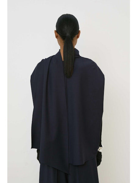 A woman with slicked-back hair and a neutral expression wears the MEIMEIJ Draped Cape Effect Top in navy over black long sleeves, paired with a matching navy skirt, set against a plain white background.