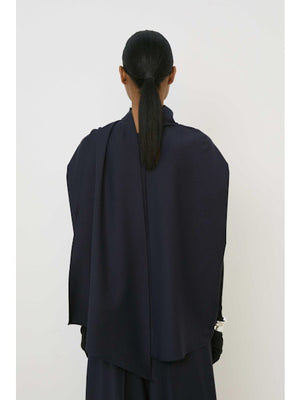 A woman with slicked-back hair and a neutral expression wears the MEIMEIJ Draped Cape Effect Top in navy over black long sleeves, paired with a matching navy skirt, set against a plain white background.