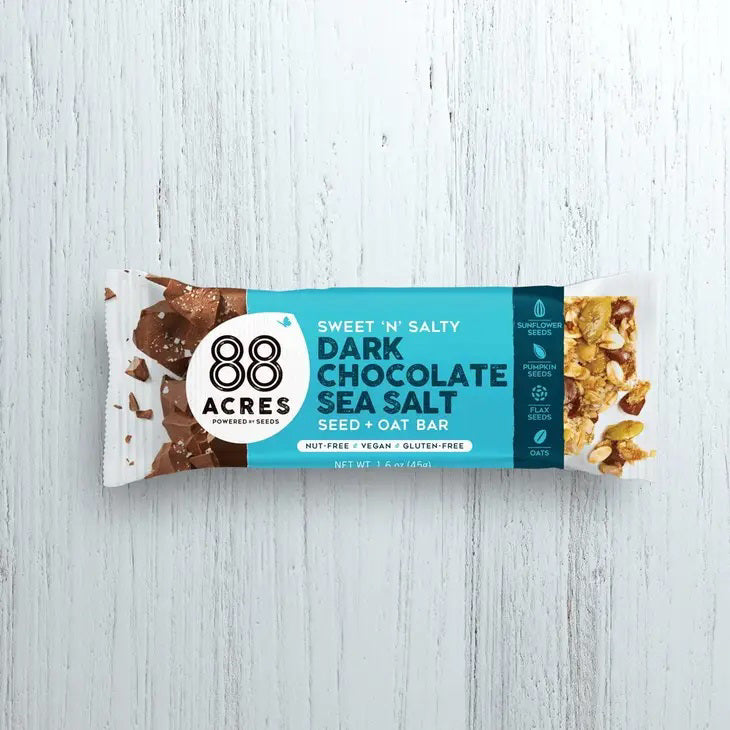 Flat lay of 88 ACRES - DARK CHOCOLATE SEA SALT SEED OAT BAR, a nut-free vegan snack, featuring opened and unopened bars in blue wrappers arranged with white boxes and chocolate pieces on a light wooden surface.