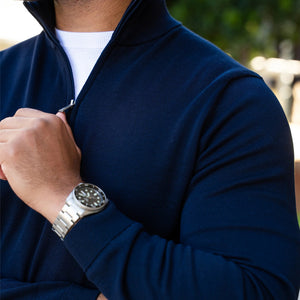 A man in the COLLARS AND CO Maranello Cotton & Silk Quarter Zip in Navy, sunglasses on, leans against a striped wall outside with one hand in his pocket and a modern glass building behind him, capturing elevated casual style.