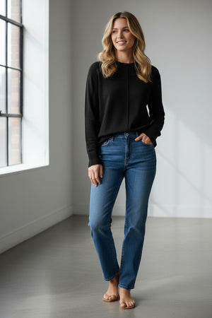 A person with long, light brown hair is wearing the KINROSS CASHMERE - HI LOW SEAMED CREW NECK CASHMERE SWEATER IN BLACK by KINROSS, styled in a relaxed classic fit with blue jeans. They are standing against a plain white background, facing the camera and smiling slightly.