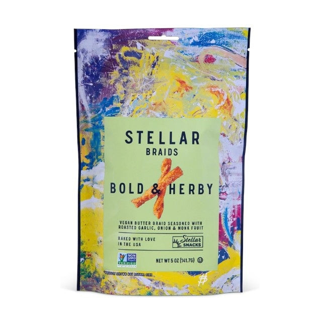 A bowl of STELLAR SNACKS - BOLD AND HERBY PRETZEL BRAIDS rests on a wooden board beside a yellow bag labeled with the STELLAR SNACKS brand name. Some pretzel braids, featuring their roasted garlic and onion flavor, are scattered on the board, while a beige cloth is partially visible.