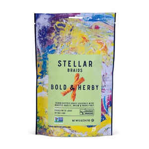 A bowl of STELLAR SNACKS - BOLD AND HERBY PRETZEL BRAIDS rests on a wooden board beside a yellow bag labeled with the STELLAR SNACKS brand name. Some pretzel braids, featuring their roasted garlic and onion flavor, are scattered on the board, while a beige cloth is partially visible.