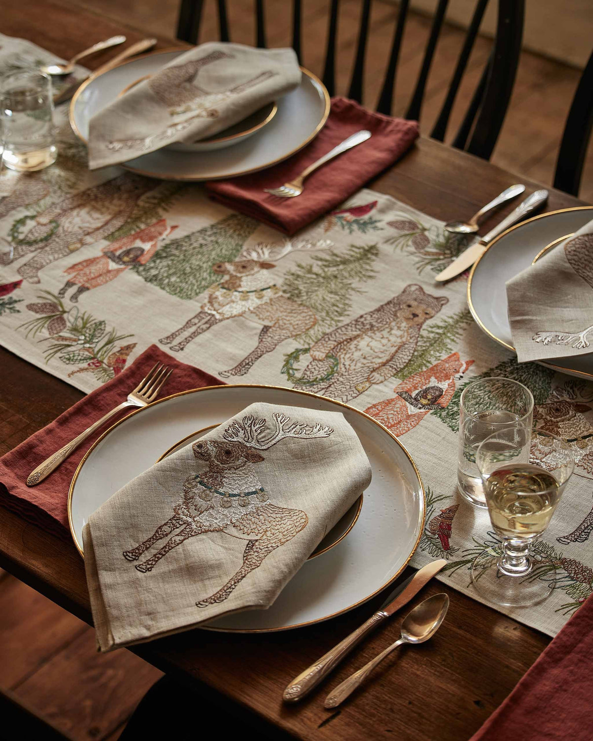 A CHRISTMAS CHEER EMBROIDERED LINEN TABLE RUNNER featuring woodland animals in winter attire and Christmas trees, draped over a wooden dining table set with plates, glassware, and candlesticks.